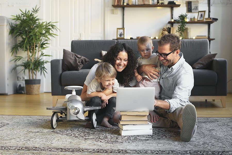 Client Center - Happy Family With Their Two Small Children Sitting on the Floor of Their Living Room Looking at a Laptop While it Rests on Top of Some Books