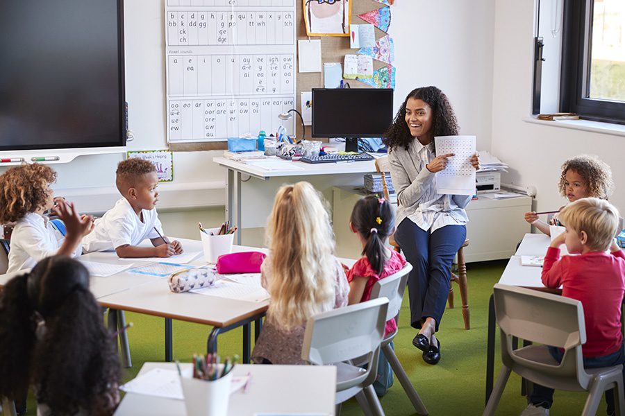 Specialized Business Insurance - Smiling Female Elementary School Teacher Sitting on a Chair Facing Her Class in a Classroom Holding up and Explaining a Worksheet to Them